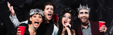 Cadılar Bayramı makyajlı bir adam siyahlar ve afişlerde çok ırklı arkadaşlarıyla karaoke yaparken el kol hareketi yapıyor.