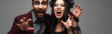 creepy interracial couple in halloween costumes showing scary gesture and growling isolated on grey, banner clipart