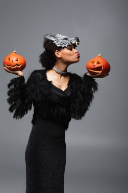 stylish african american woman in wolf halloween mask holding carved pumpkins and blowing air kiss isolated on grey clipart