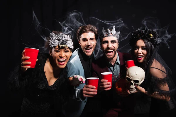 Amigos Multiétnicos Disfraces Halloween Sosteniendo Copas Plástico Riendo Cámara Negro —  Fotos de Stock