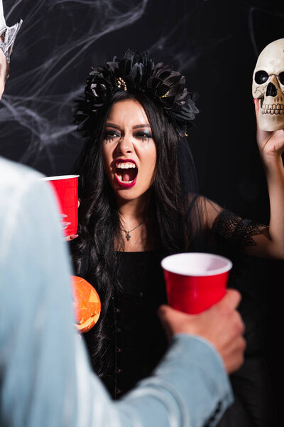 asian woman in vampire costume growling while frightening blurred man with spooky skull on black