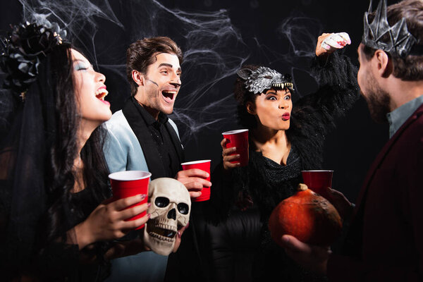 african american woman in wolf mask pouting lips while frightening multiethnic friends with toy hand on black 