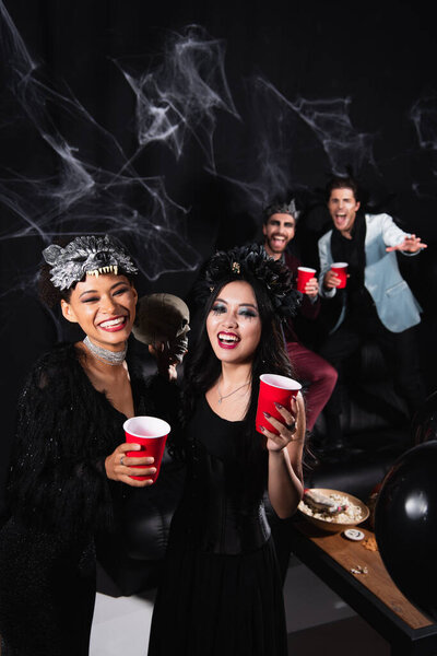 happy interracial women in halloween costumes near man waving hand on blurred and black background