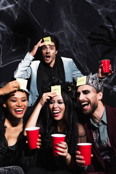 изумленный мужчина смотрит в камеру рядом с многонациональными друзьями, играющими в угадай, кто играет во время Хэллоуинской вечеринки на черном