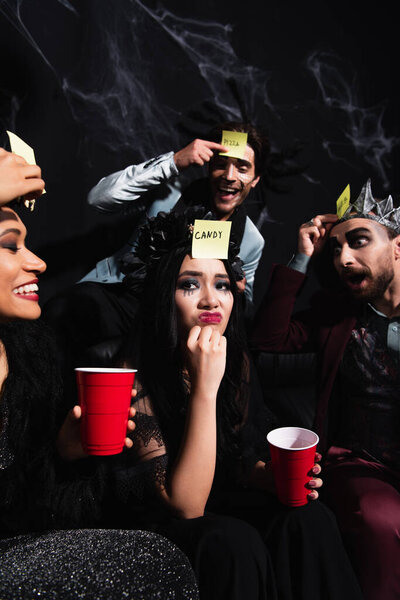 вдумчивая азиатка возле взволнованных мультикультурных друзей, играющих в угадай, кто играет во время Хэллоуинской вечеринки на черном