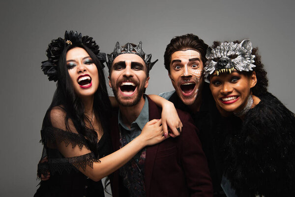 cheerful african american woman hugging man in vampire king crown during halloween party with friends isolated on grey