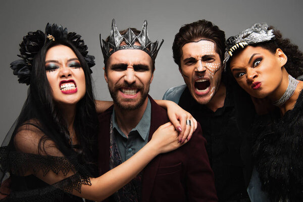 multiethnic friends in scary halloween costumes grimacing at camera isolated on grey