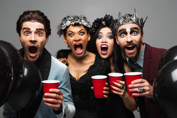 spooky multiethnic friends with plastic cups screaming and growling at camera on halloween party isolated on grey