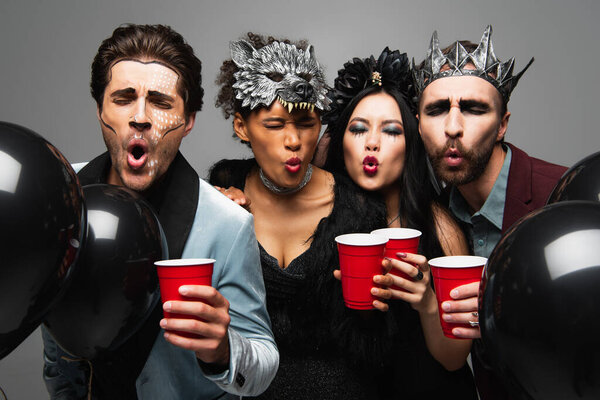 multiethnic friends in halloween costumes blowing at hot beverage in plastic cups isolated on grey