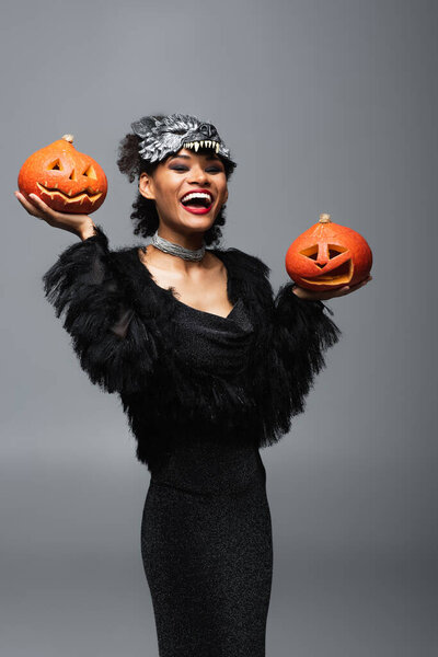 excited african american woman in black dress and wolf mask holding halloween pumpkins isolated on grey
