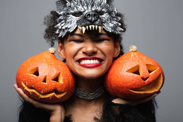 overjoyed african american woman in wolf halloween mask holding carved pumpkins and smiling with closed eyes isolated on grey