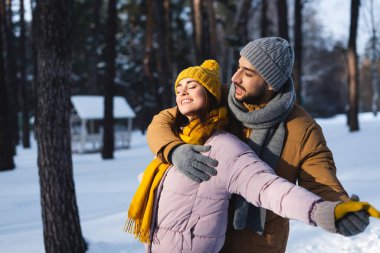 Man hugging and holding hand of happy girlfriend with closed eyes in winter park  clipart