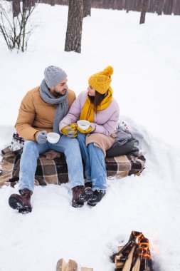Kar parkında şenlik ateşinin yanında el ele tutuşan kahve bardaklı çift. 