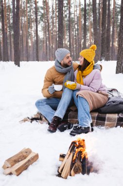 Mutlu genç çift ellerinde şenlik ateşinin yanında kahve ve kış parkında kütükler tutuyor. 