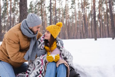 Kış parkında örgü şapkalı ve battaniyeli güzel kız arkadaşına sarılan mutlu bir adam. 