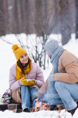 Elinde fincanla gülümseyen kadın kış parkında ateşin yanında sevgilisine bakıyor. 