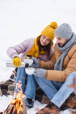 Bir adam, kız arkadaşının yanında termoslu bardak tutuyor ve kış parkında şenlik ateşini bulanıklaştırıyor. 