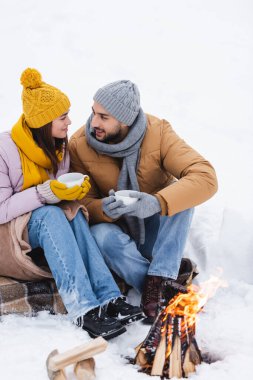 Eldivenli gülümseyen adam kız arkadaşının yanında bardak tutuyor ve karda ateş yakıyor. 