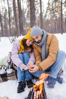 Kış kıyafetli genç çift parktaki şenlik ateşinin yanında ellerini uyarıyor. 