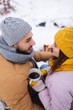 Kış parkında gülümseyen erkek arkadaşın yanında elinde kahve fincanı tutan örgü şapkalı kadının yüksek açılı görüntüsü. 