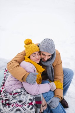 Parkta karlı havada, battaniyeli gülümseyen bir kadının sevgilisine sarıldığı yüksek açı. 