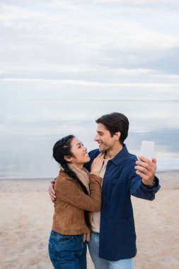 Gülümseyen ırklar arası bir çift sahilde sarılıyor ve selfie çekiyor. 