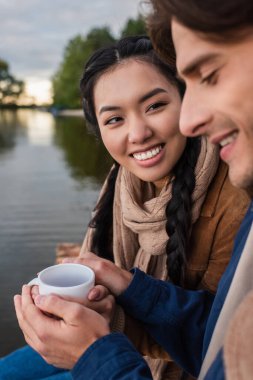 Gülümseyen Asyalı kadın bardağını açık havada ve bulanık erkek arkadaşının yanında tutuyor. 
