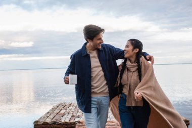 Elinde kupayla gülümseyen adam iskelede battaniyeli Asyalı kız arkadaşına sarılıyor. 