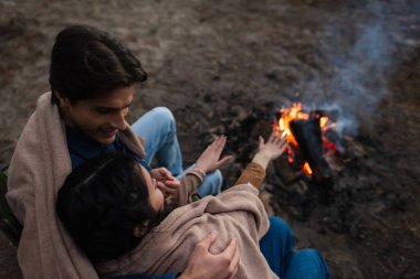 Bulanık kamp ateşinin yanında battaniyeli Asyalı kız arkadaşına sarılan adamın tepeden bakışı 