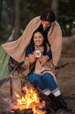Bulanık kamp ateşinin yanında elinde fincanla Asyalı kız arkadaşının elini tutan bir adam. 