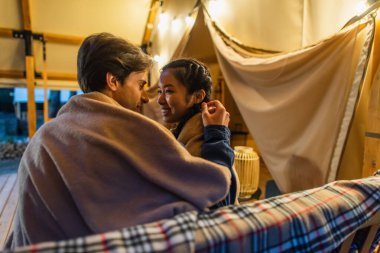 Battaniyeli bir adam dışarıdaki glamping evinin yanındaki bankta gülümseyen Asyalı kız arkadaşına dokunuyor. 