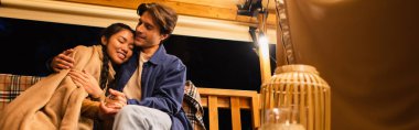 Irklar arası çift, glamping evinin terasında, ışık çelengi yakınında bankta el ele tutuşuyorlar. 