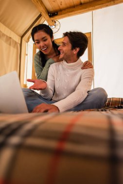 Gülümseyen adam Glamping House 'da Asyalı kız arkadaşının yanındaki bilgisayarı işaret ediyor. 