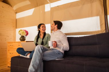 Glamping House 'daki koltukta dizüstü bilgisayar kullanan çok ırklı neşeli bir çift. 