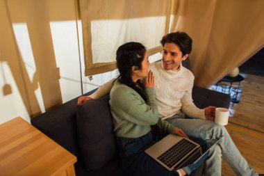 Dizüstü bilgisayarı olan Asyalı bir kadın Glamping House 'da elinde kupayla sevgilisiyle konuşuyor. 
