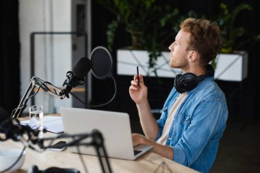 Düşünceli radyo sunucusu kulaklıklarla stüdyoda cihazların yanında kalem ve mikrofon tutuyor. 