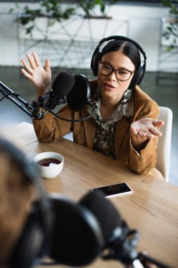 Asyalı radyo sunucusunun gözlüklü ve kulaklıklı yüksek açılı görüntüsü bir fincan kahvenin yanında el kol hareketi ve podcast sırasında bulanık iş arkadaşı