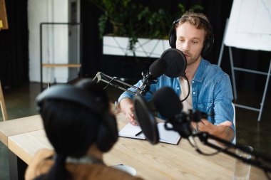 Kulaklıkla radyo sunucusu mikrofonla konuşuyor ve podcast sırasında bulanık meslektaşına bakıyor.