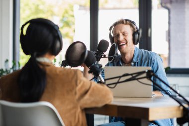 Kulaklıkla gülümseyen radyo sunucusu mikrofonla konuşuyor ve podcast sırasında bulanık meslektaşına bakıyor.
