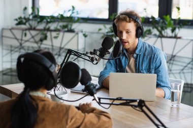 Kulaklıklı radyo sunucusu dizüstü bilgisayarın yanında mikrofonla konuşuyor ve podcast sırasında meslektaşı bulanıklaşıyor