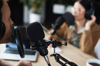 radio host in wireless headphones talking in microphone near blurred asian colleague  clipart