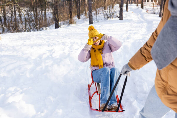 Smiling woman on sled showing love sign at blurred boyfriend in winter park 
