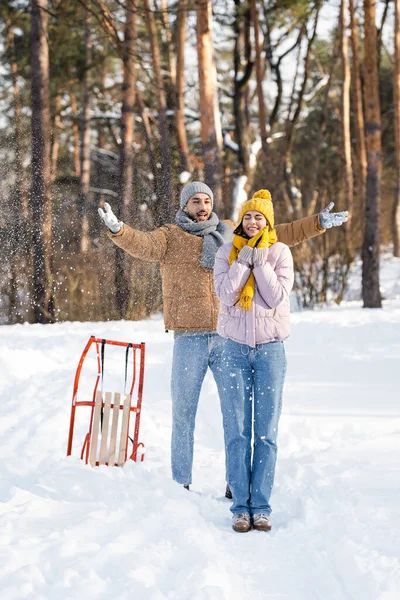 stock image Cheerful man throwing snow near girlfriend with closed eyes and sled in winter park 