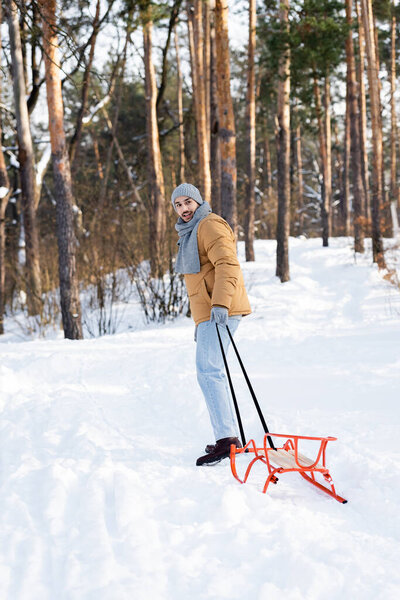 Man in hat and scarf smiling at camera while pulling sleigh in winter park 