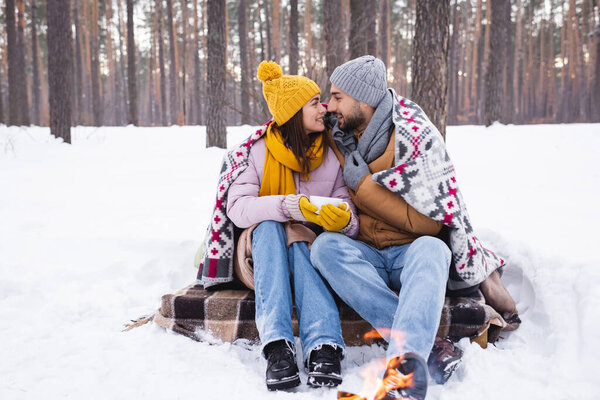 Side view of positive couple with cup and blanket looking at each other near blurred bonfire in winter park 