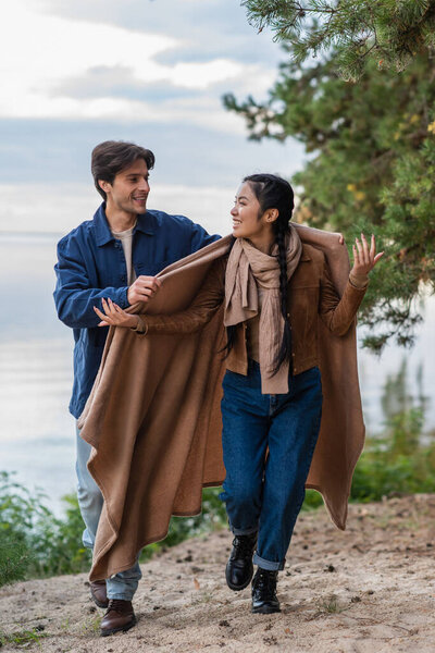 Smiling man holding blanket near asian girlfriend walking on beach 