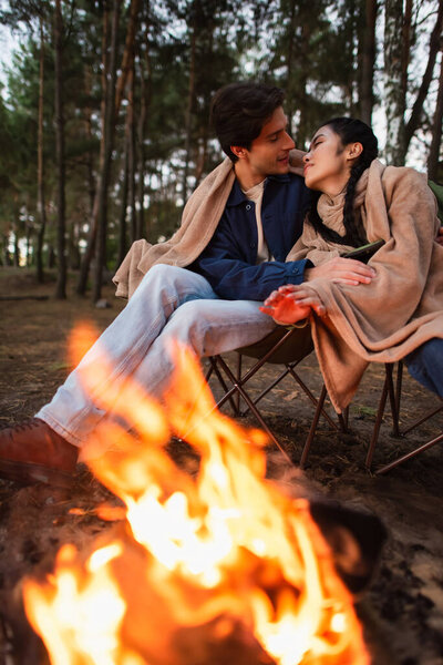 Interracial couple in blankets kissing near blurred campfire 