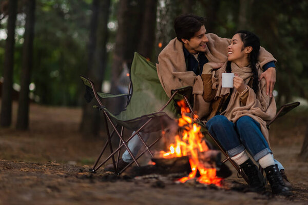 Multiethnic couple in blankets with cup taking near blurred campfire 