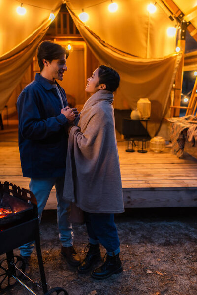 Вид сбоку межрасовой пары, держащейся за руки возле гриля и пылающего дома в вечернее время 