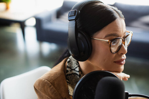 asian radio host in glasses and headphones looking away during broadcast 
